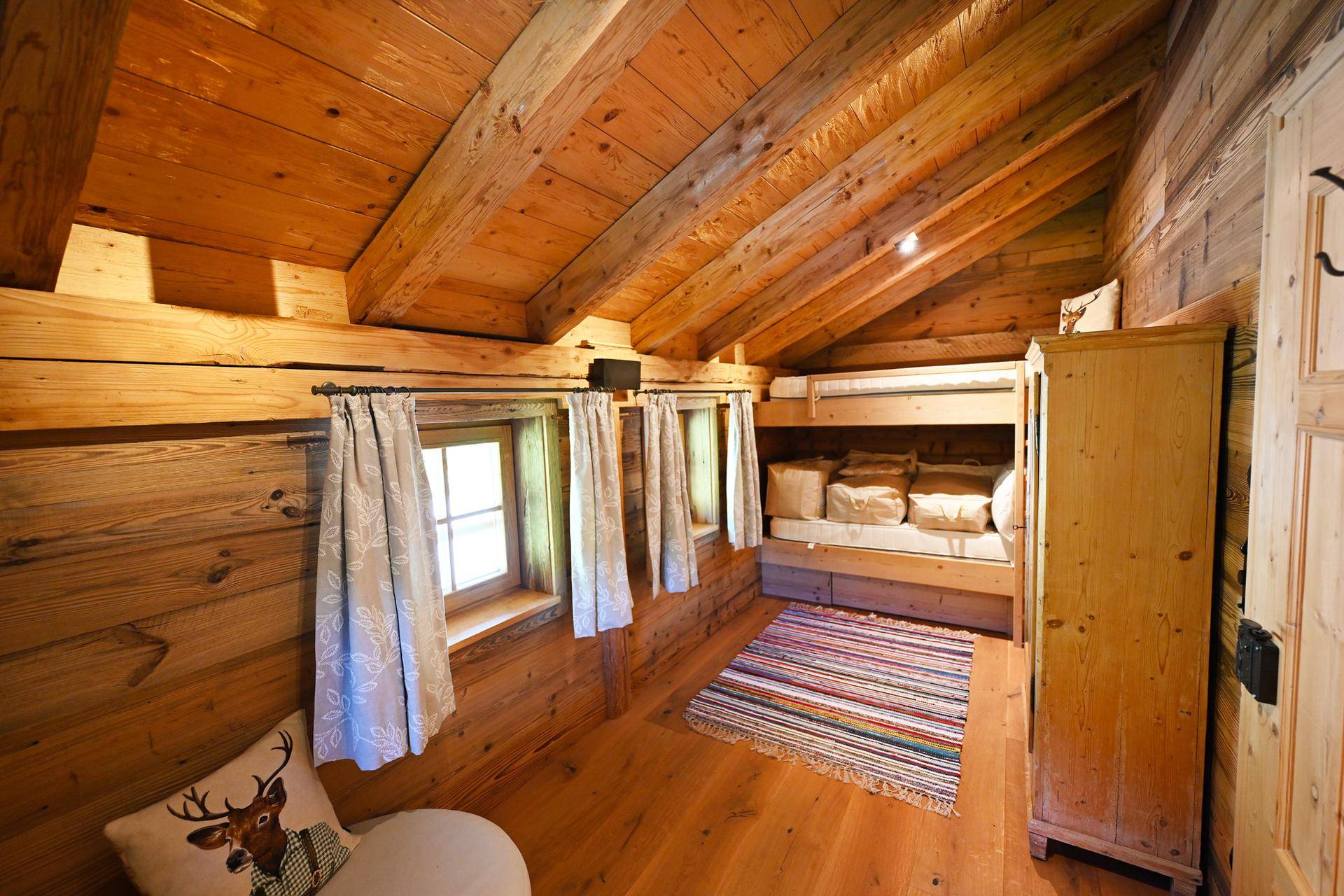 Rustikales Schlafzimmer mit Etagenbetten, Holzwänden und Holzbalkendecke, ergänzt durch einen farbenfrohen Teppich.
