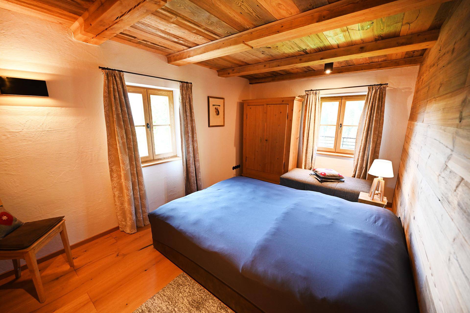 Helles Schlafzimmer mit Doppelbett, weißen Wänden, Holzbalkendecke und zwei Fenstern für viel Tageslicht.