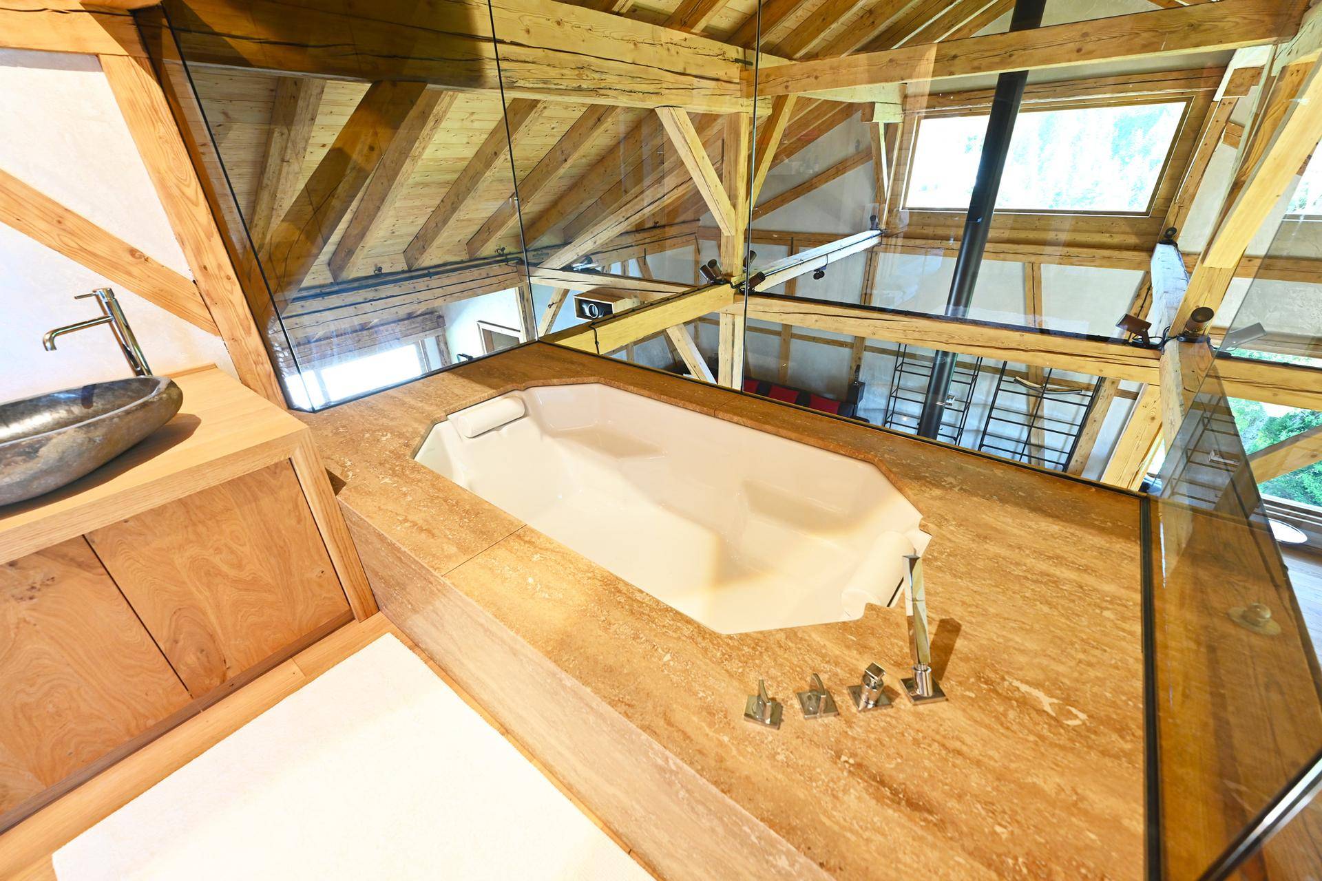 Elegante Badewanne mit Natursteinumrandung und Glasabtrennung unter einer beeindruckenden Holzbalkendecke.