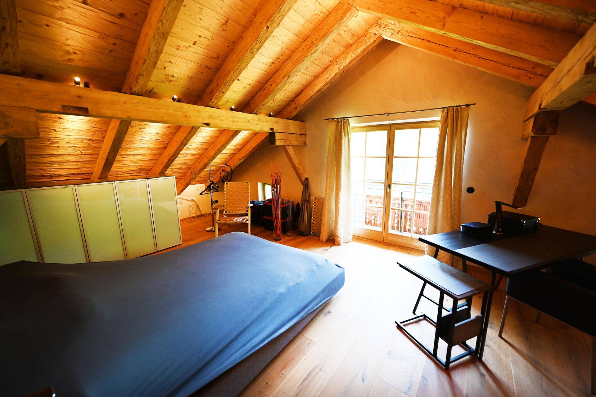 Helles Schlafzimmer mit Doppelbett, Holzbalkendecke und großem Fenster mit Zugang zu einem Balkon.