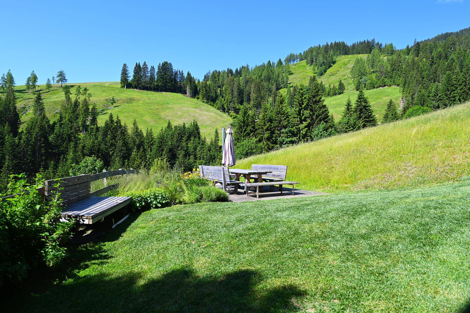 Gepflegter Gartenbereich mit rustikalen Holzmöbeln und Sonnenschirm, umgeben von grünen Hügeln.