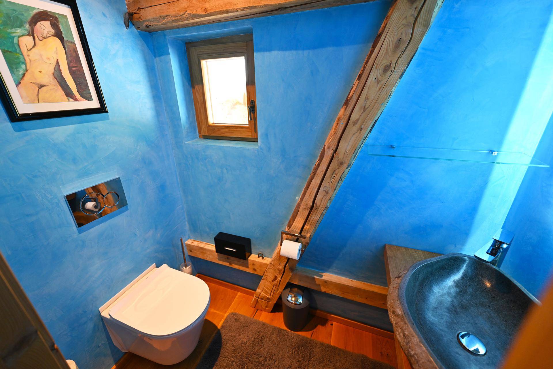 Modernes Gäste-WC mit blauen Wänden, Holzbalken und wandhängendem WC, bietet ein Fenster für Belüftung.