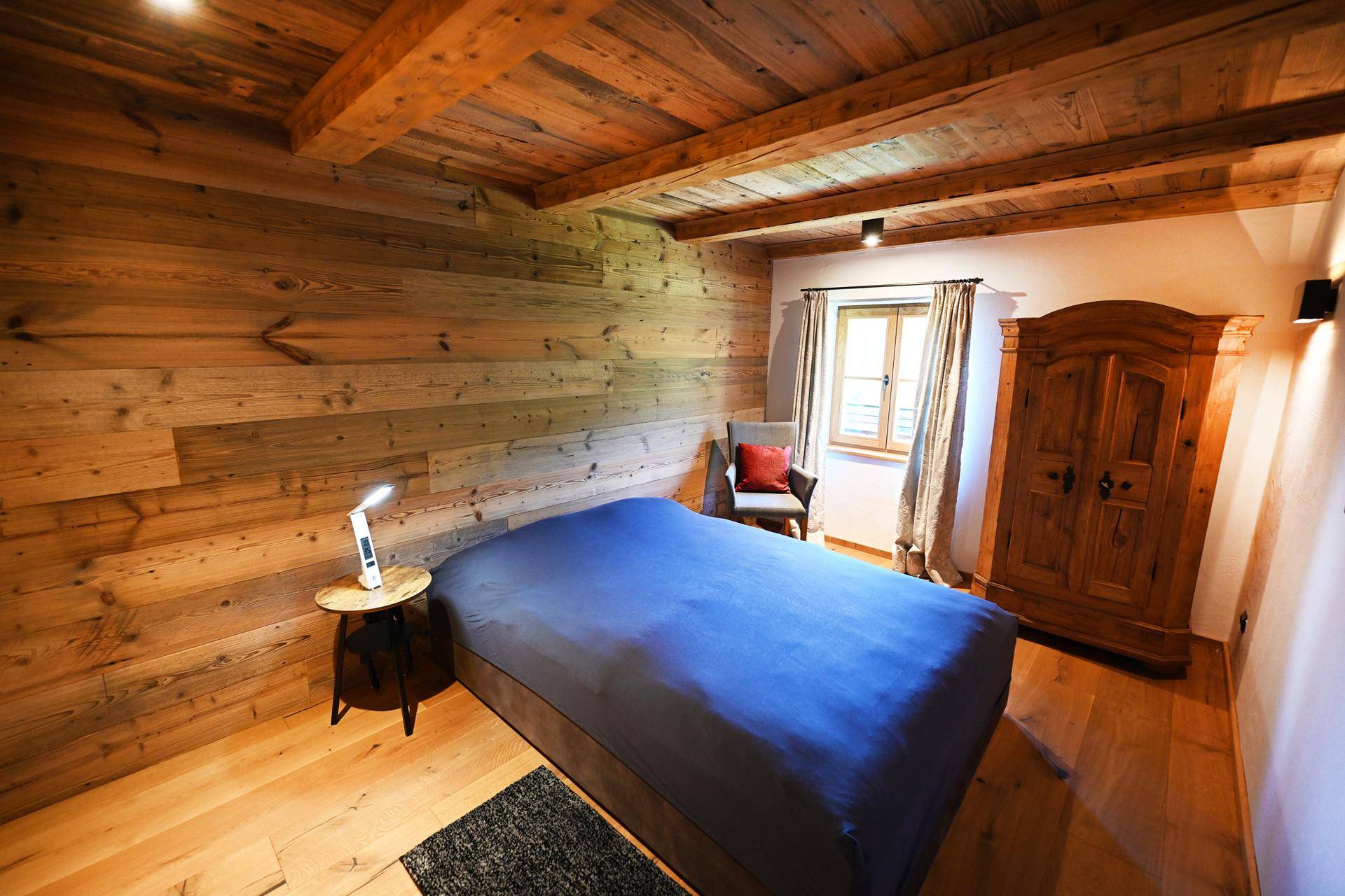 Komfortables Schlafzimmer mit Doppelbett, Holzwand, Holzbalkendecke und einem traditionellen Holzschrank.