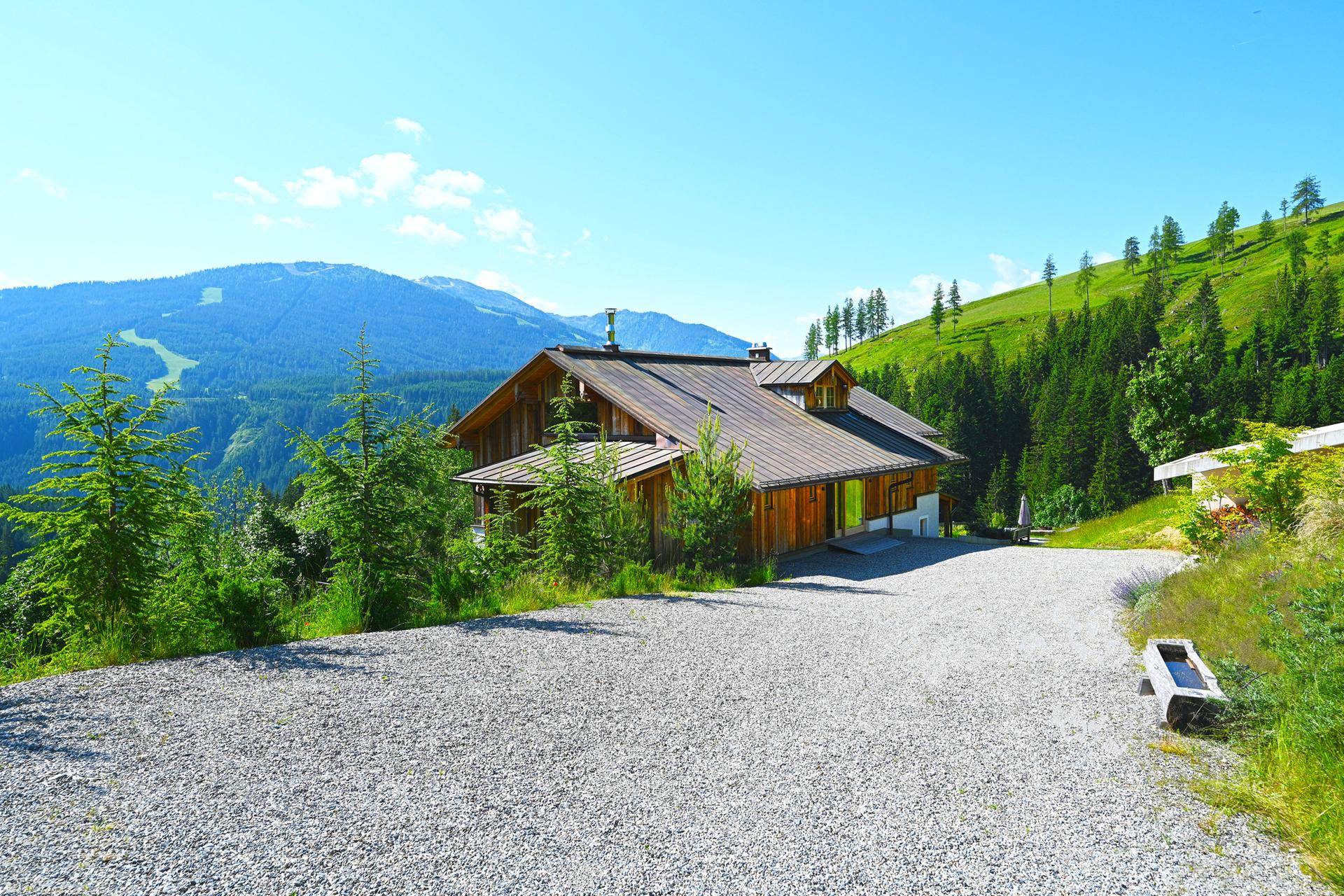 Chalet mit großem Kiesparkplatz und Panoramablick auf die umliegenden Berge und Wälder.