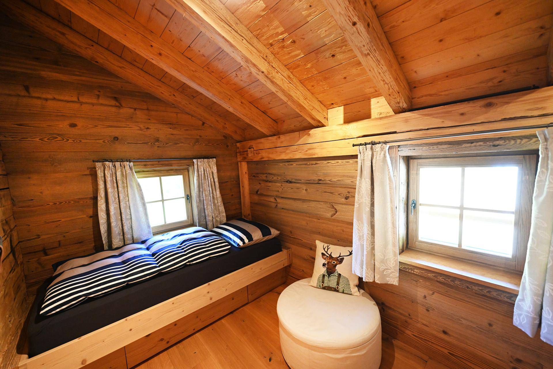 Gemütliches Schlafzimmer im alpinen Stil mit Holzwänden, Holzbalkendecke und zwei Fenstern für natürliche Helligkeit.