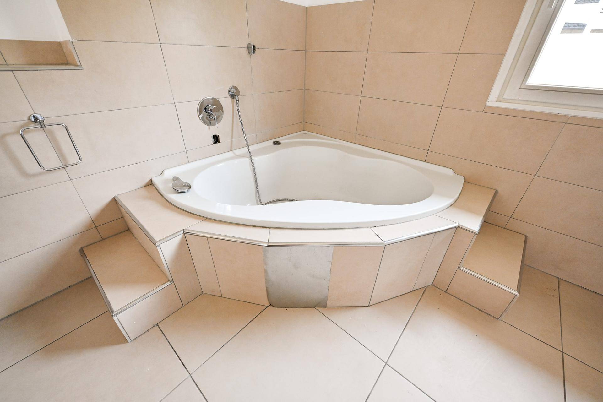 Eckbadewanne mit Fliesenverkleidung und Duscharmatur in hellem Badezimmer.