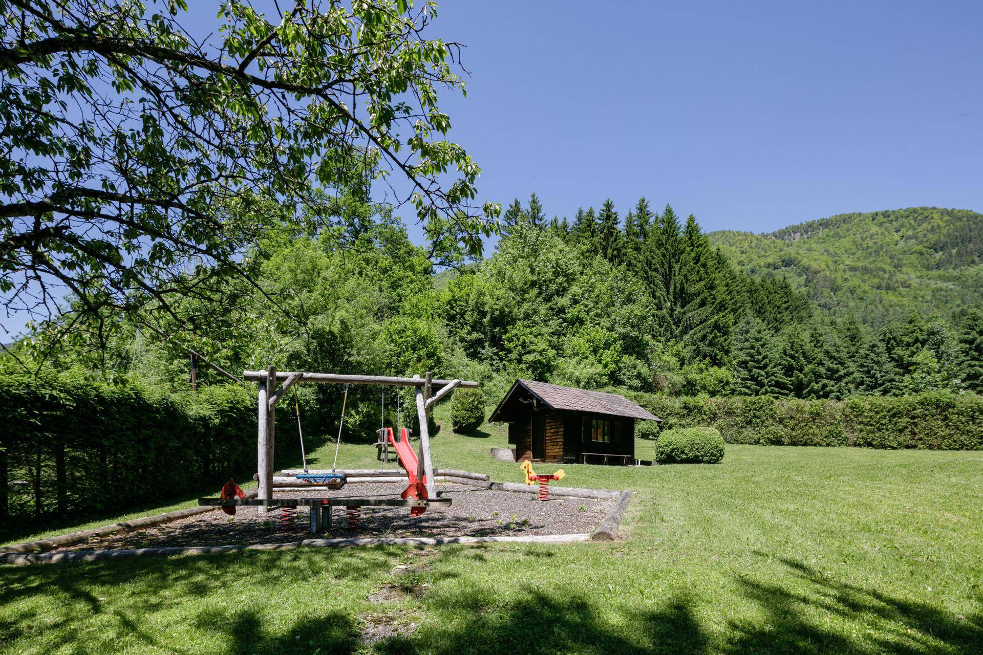 Großer Gartenbereich mit Kinderspielplatz, Schaukeln und Rutsche, umgeben von Bäumen und Hügeln.