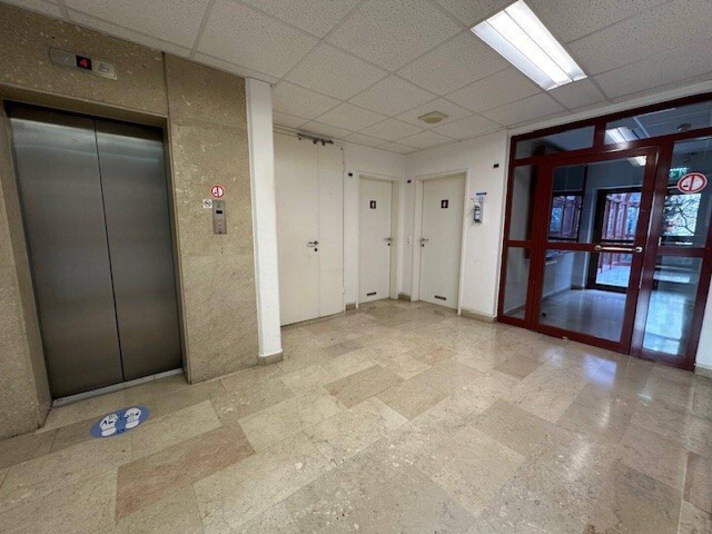 Eingangsbereich mit Aufzug, mehreren Türen und hellem Fliesenboden.