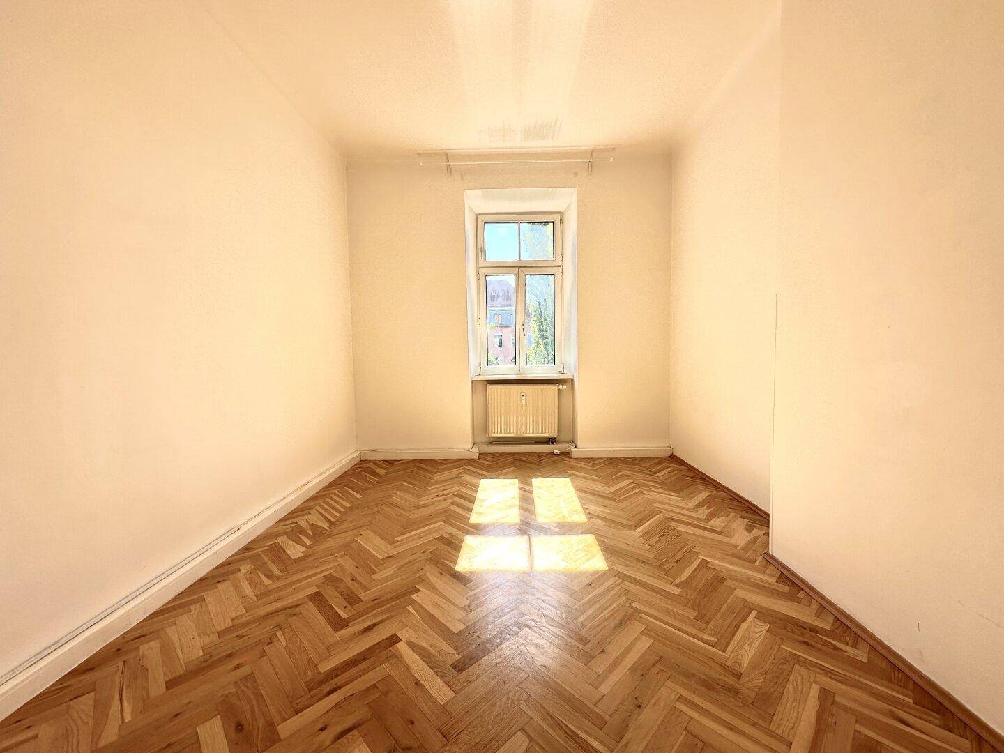 Helles Zimmer mit Fischgrätparkettboden, beigen Wänden und einem Fenster, das Sonnenlicht hereinlässt.