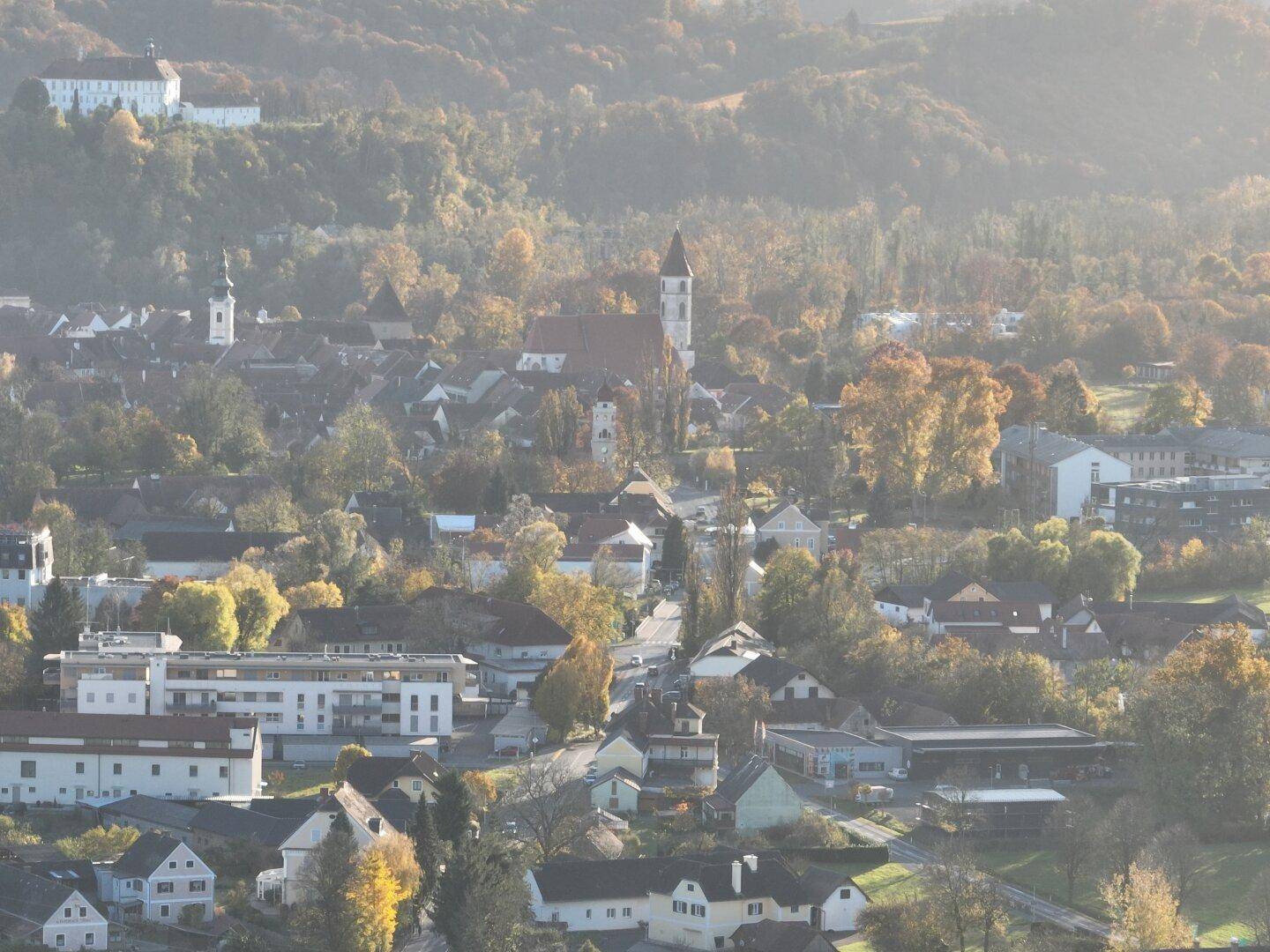 Dorfansicht in hügeliger Landschaft mit historischen Gebäuden und herbstlicher Baumkulisse im Dunst.