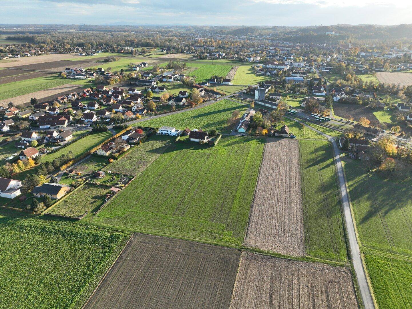 Luftaufnahme einer weitläufigen Wohnsiedlung umgeben von grünen Feldern und Ackerland bei sonnigem Wetter.