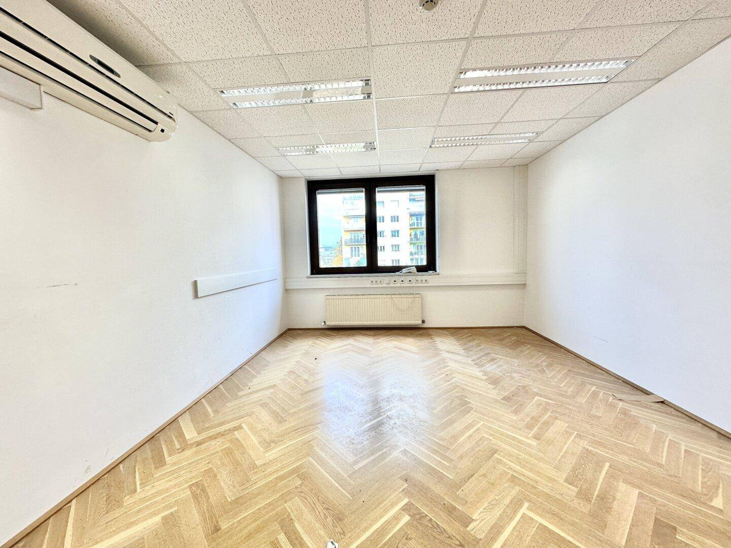 Leeres Büro mit Fischgrätparkettboden, einem Fenster und einer Klimaanlage an der Wand.
