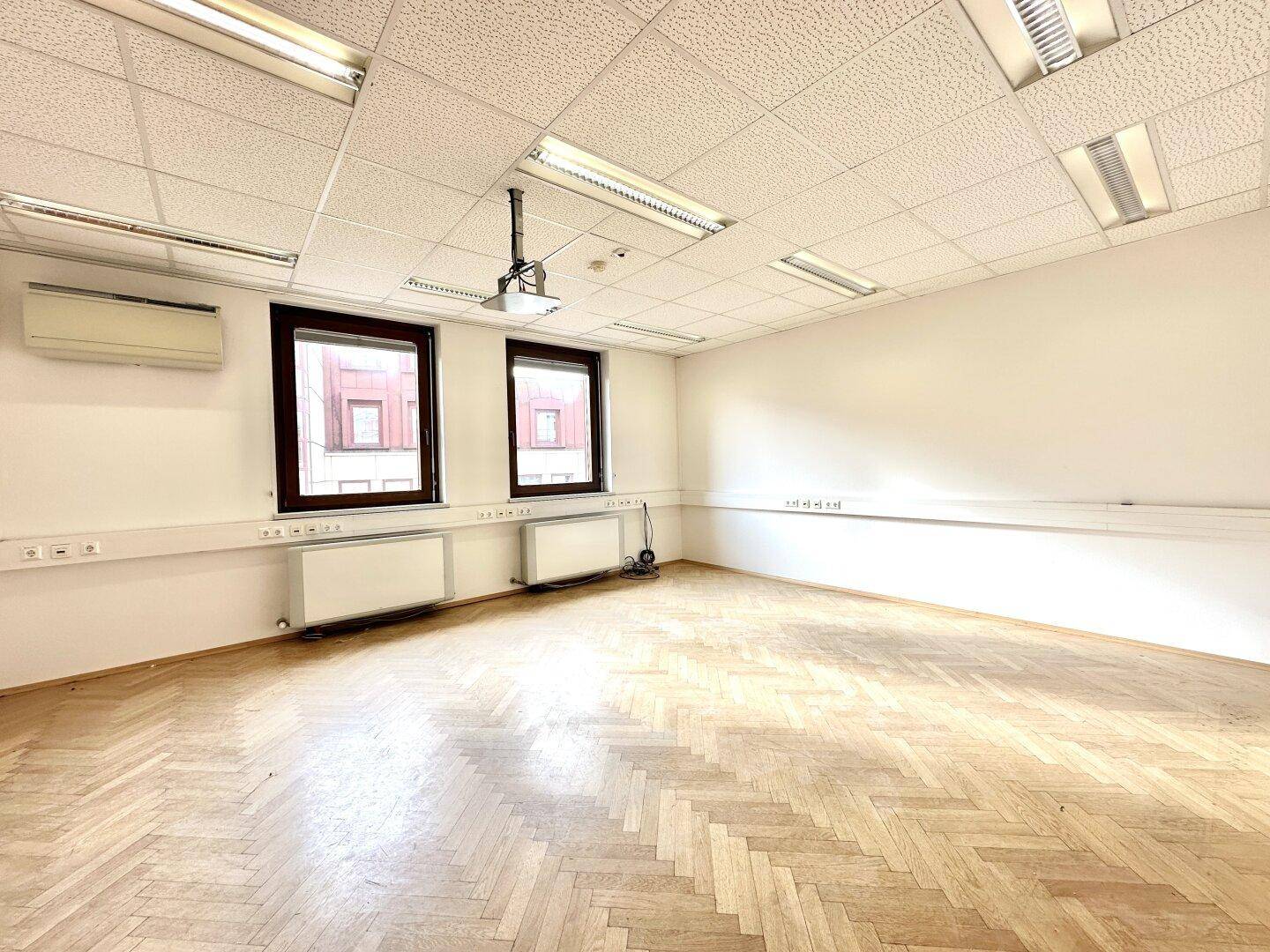 Leeres Büro mit Fischgrätparkettboden, großen Fenstern und Heizkörpern unter den Fenstern.