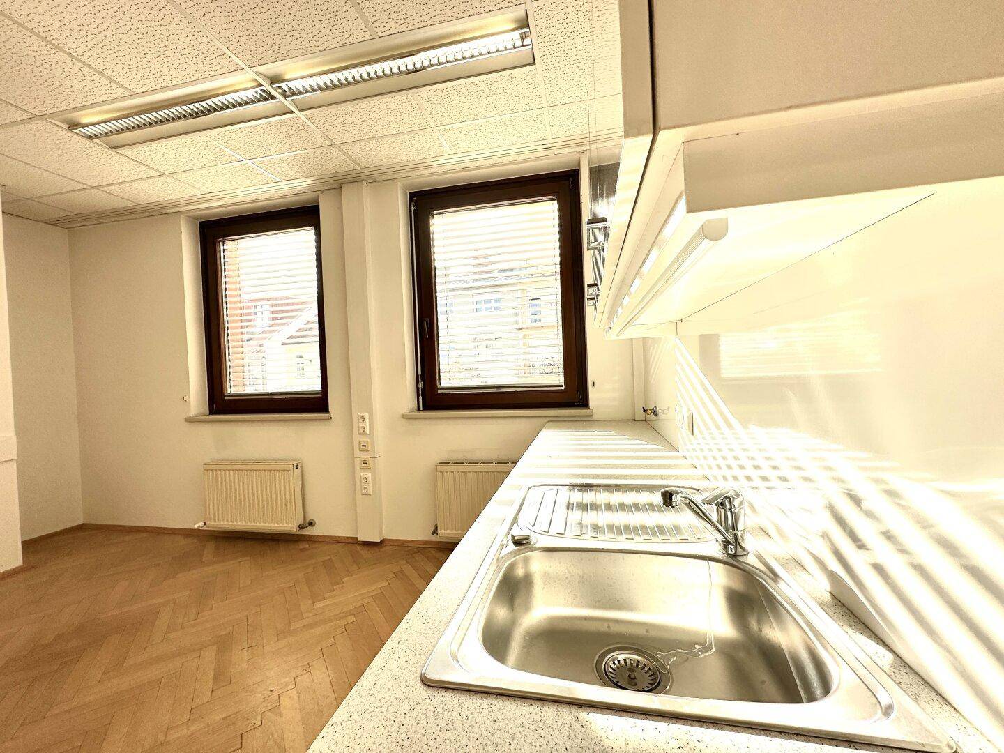 Kleine Küchenzeile mit Spüle und Arbeitsplatte, beleuchtet durch zwei Fenster.