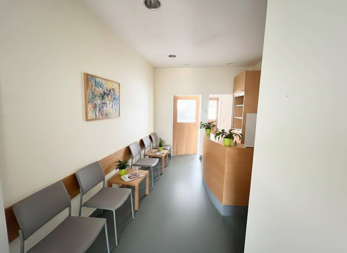Heller Wartebereich mit mehreren grauen Sitzgelegenheiten, Holztischen und einer geschwungenen Rezeption.