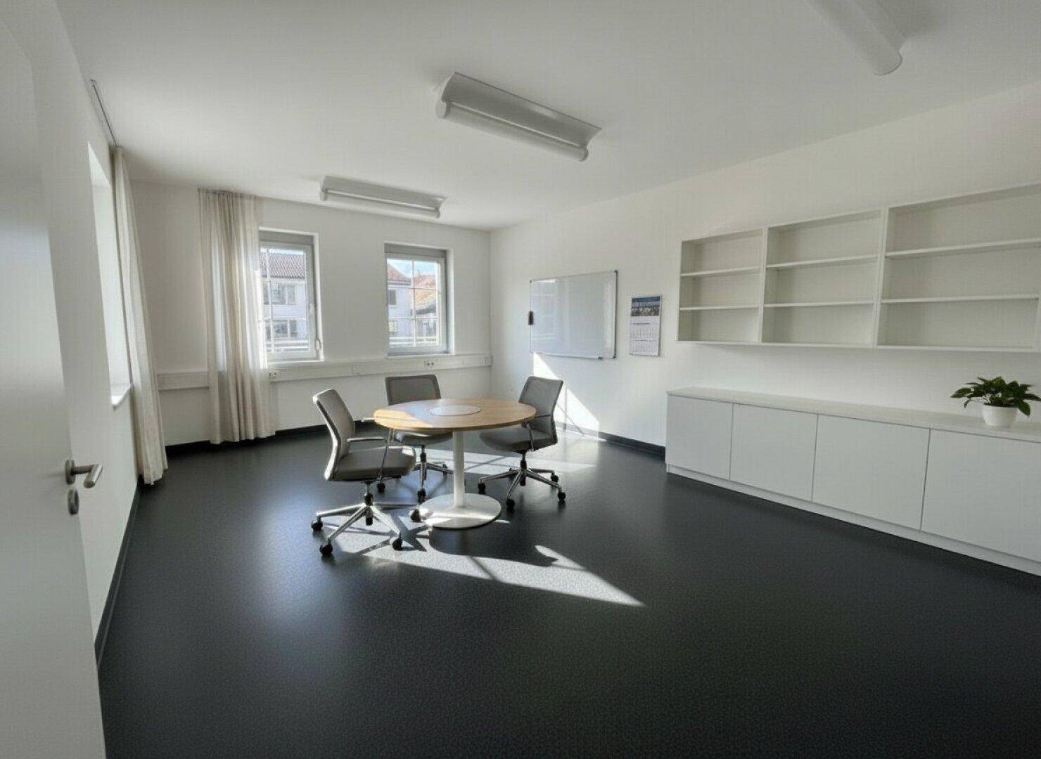 Heller Büroraum mit Besprechungstisch, Bürostühlen und weißen Regalen an der Wand, gut beleuchtet.