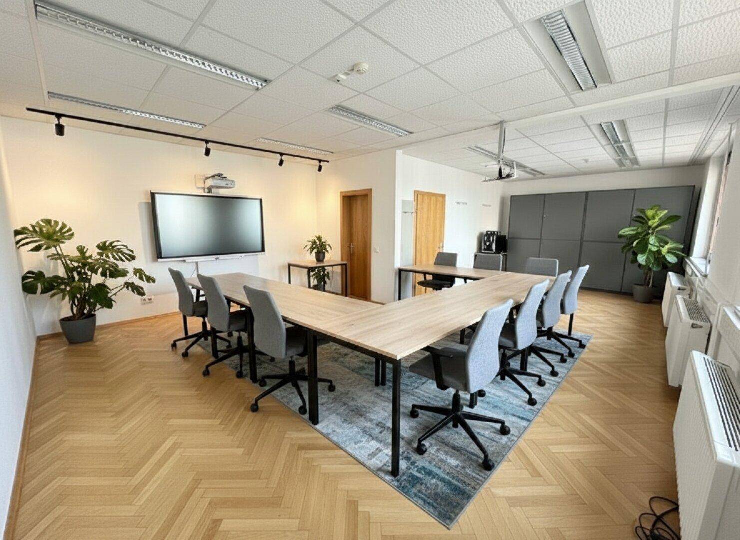 Konferenzraum mit U-förmigem Tisch, modernen Stühlen und großem Wandbildschirm für Präsentationen.