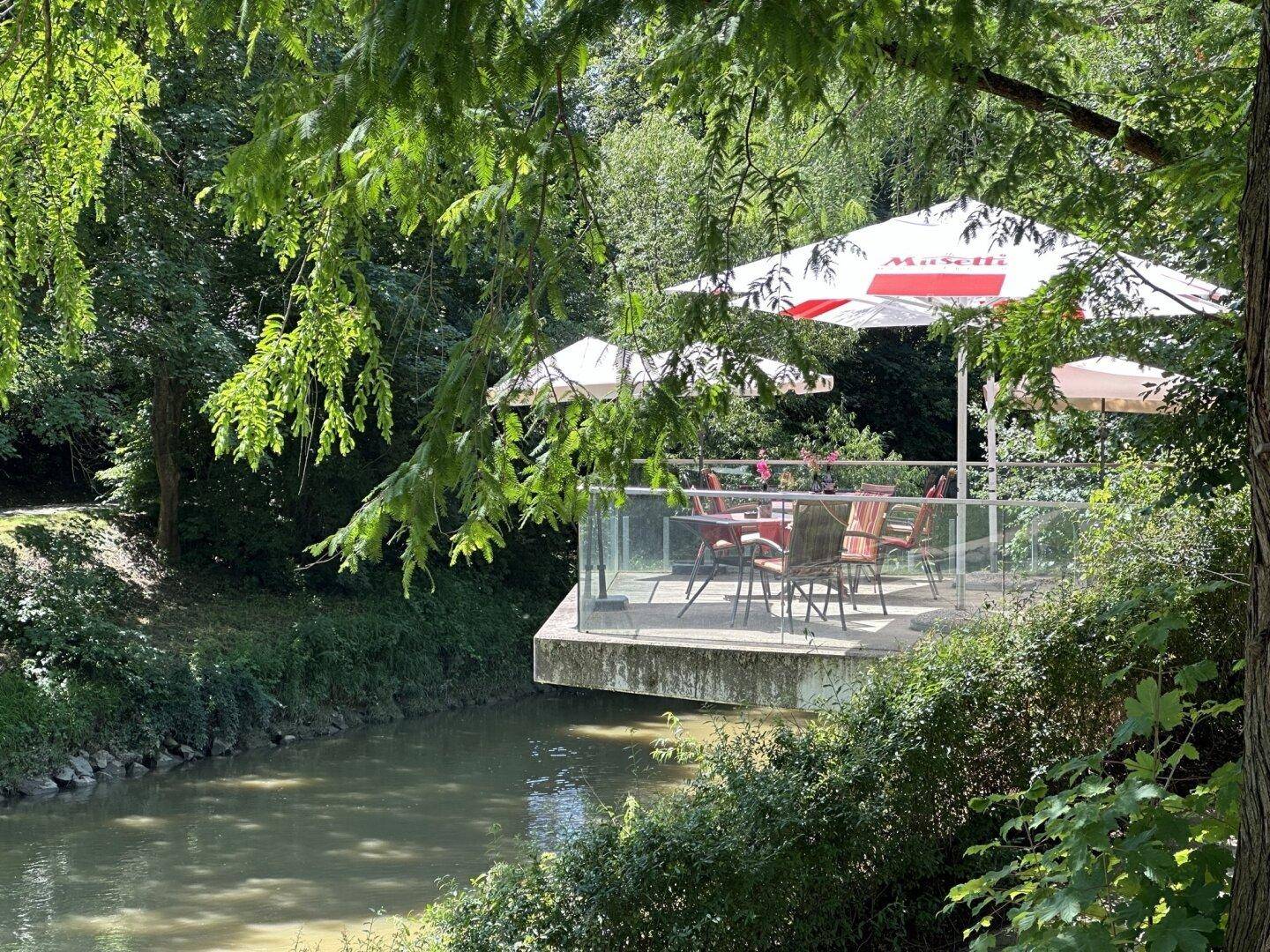 Idyllische Terrasse am Flussufer mit Sonnenschirmen und Sitzgelegenheiten, umgeben von üppigem Grün.