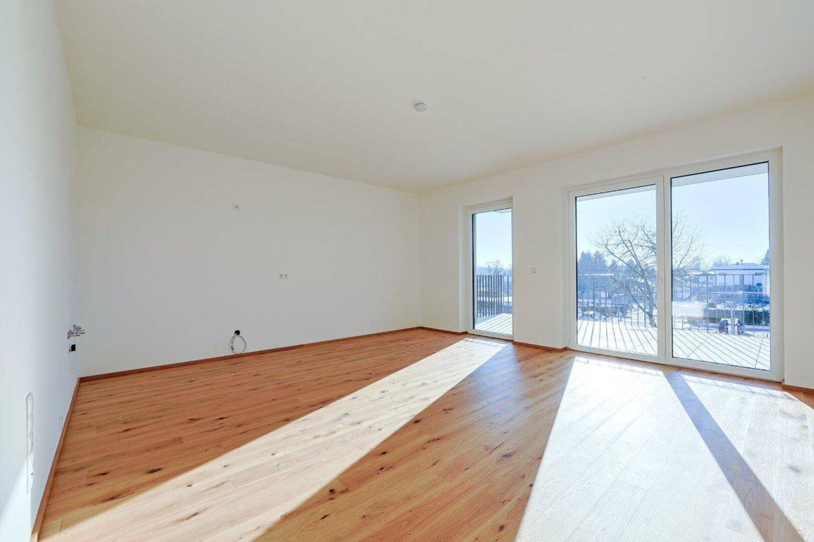 Geräumiges, leeres Wohnzimmer mit Holzboden und Zugang zu einem Balkon, durchflutet von Sonnenlicht.