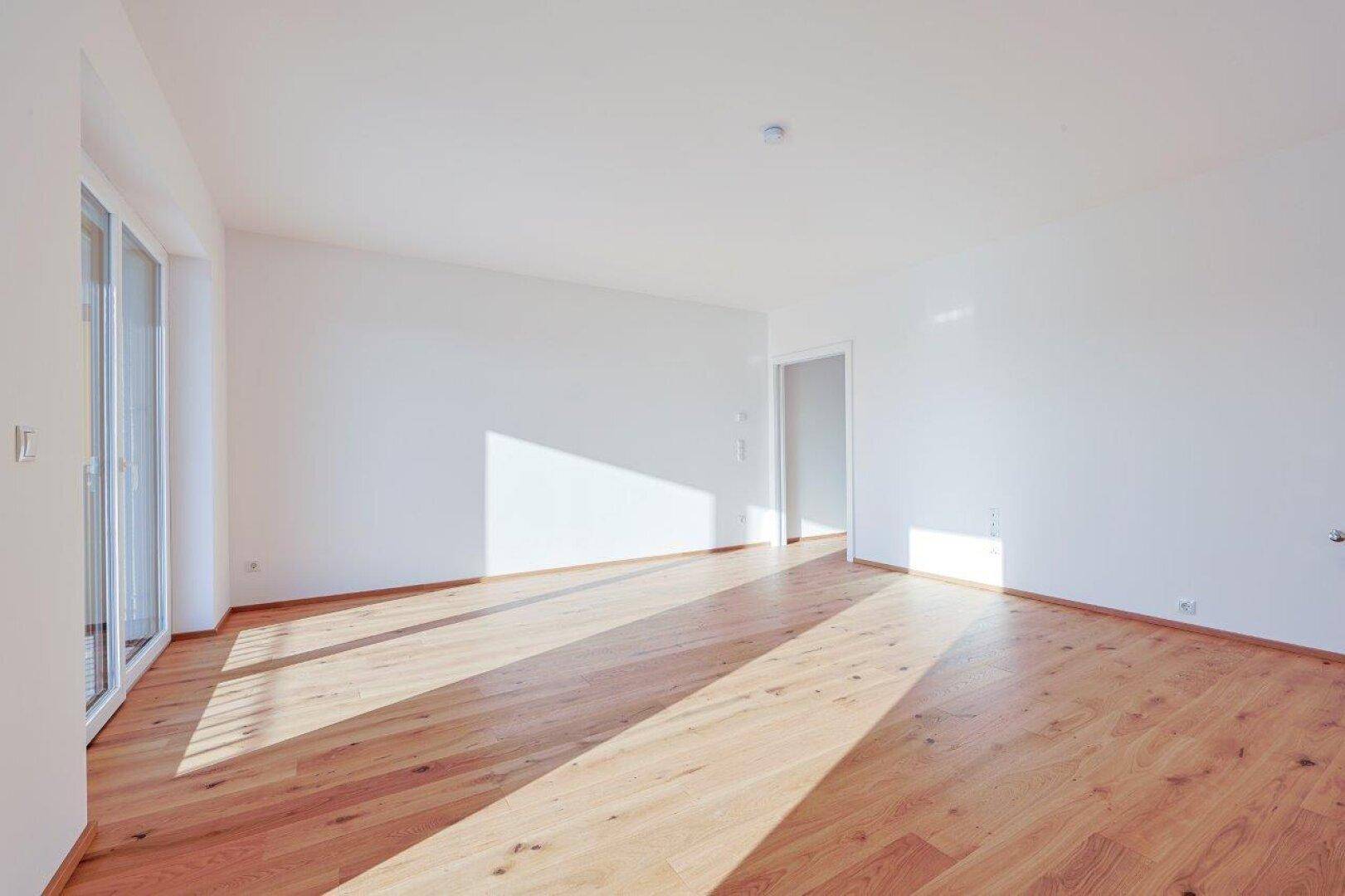 Helles, leeres Wohnzimmer mit Holzboden und Zugang zu weiteren Räumen, sonnendurchflutet.