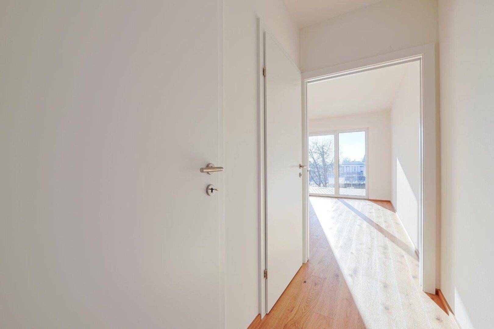 Flur mit Fliesenboden und offener Tür zu einem Zimmer mit Holzboden und Balkonzugang.