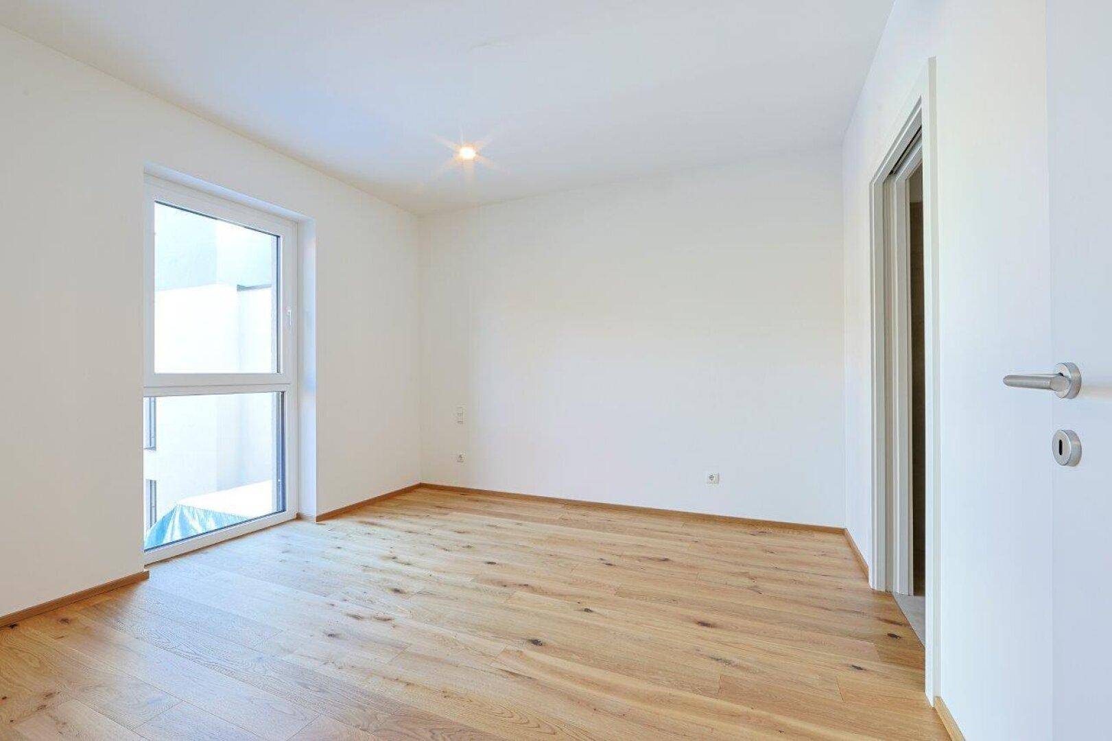 Leeres Schlafzimmer mit Holzboden und einem großen Fenster, das viel Tageslicht hereinlässt.