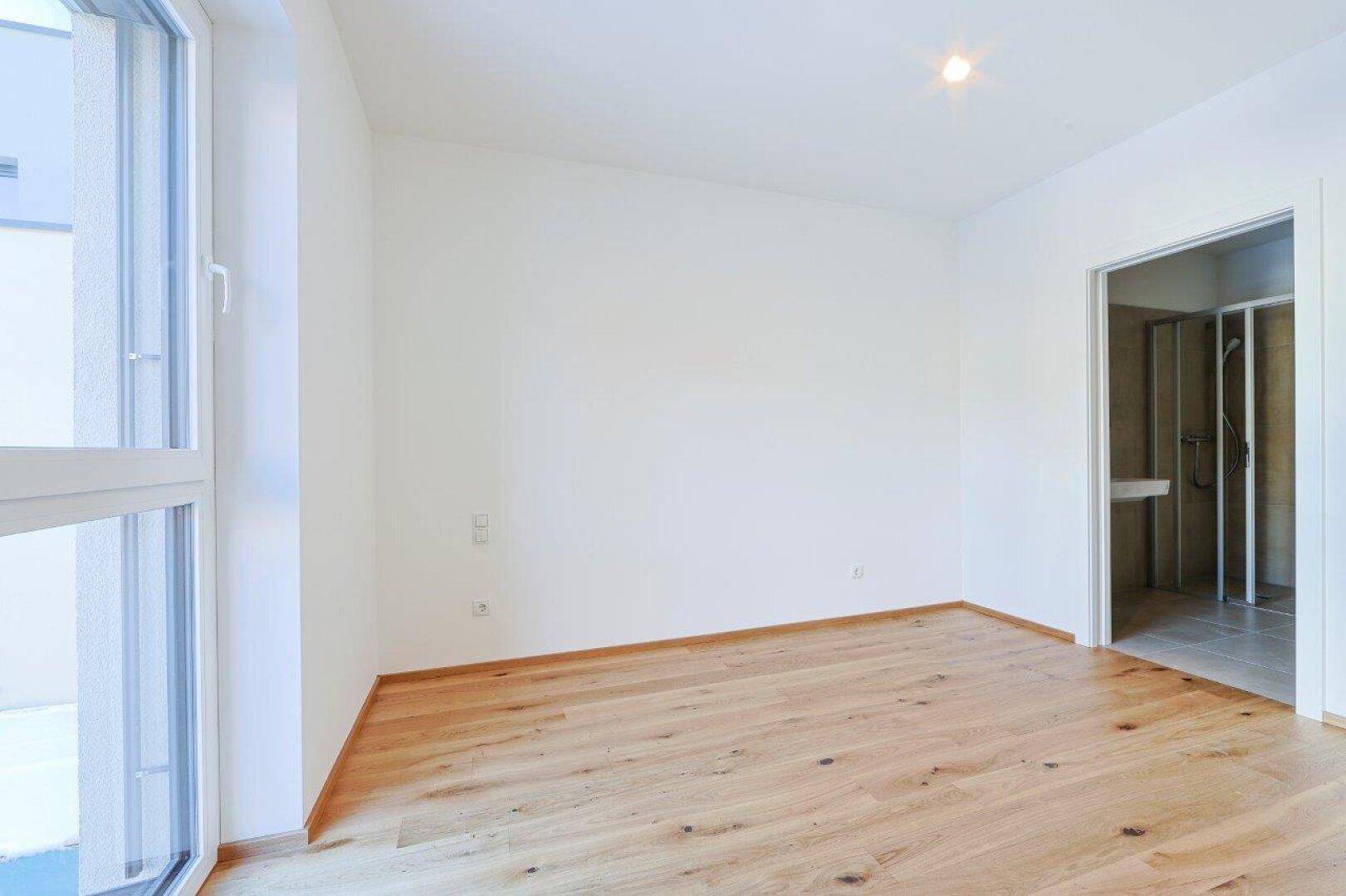 Helles Schlafzimmer mit Holzboden und Blick auf das angrenzende Badezimmer mit Dusche.