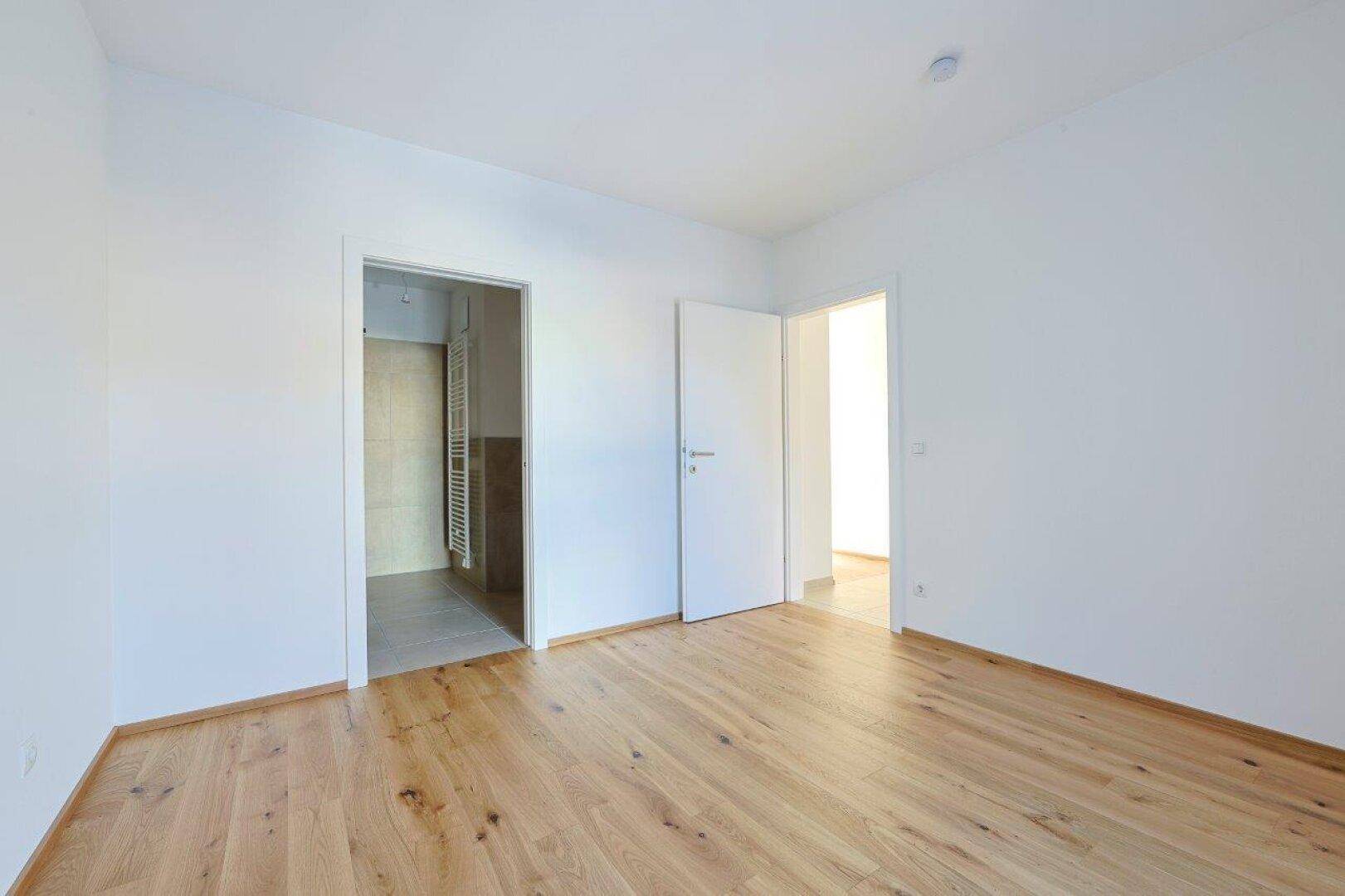 Leeres Schlafzimmer mit Holzboden und offenen Türen zu einem Badezimmer und einem weiteren Raum.