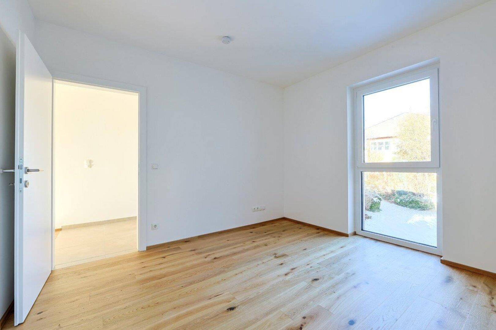 Helles Schlafzimmer mit Holzboden und großem Fenster, das einen Blick ins Grüne bietet.