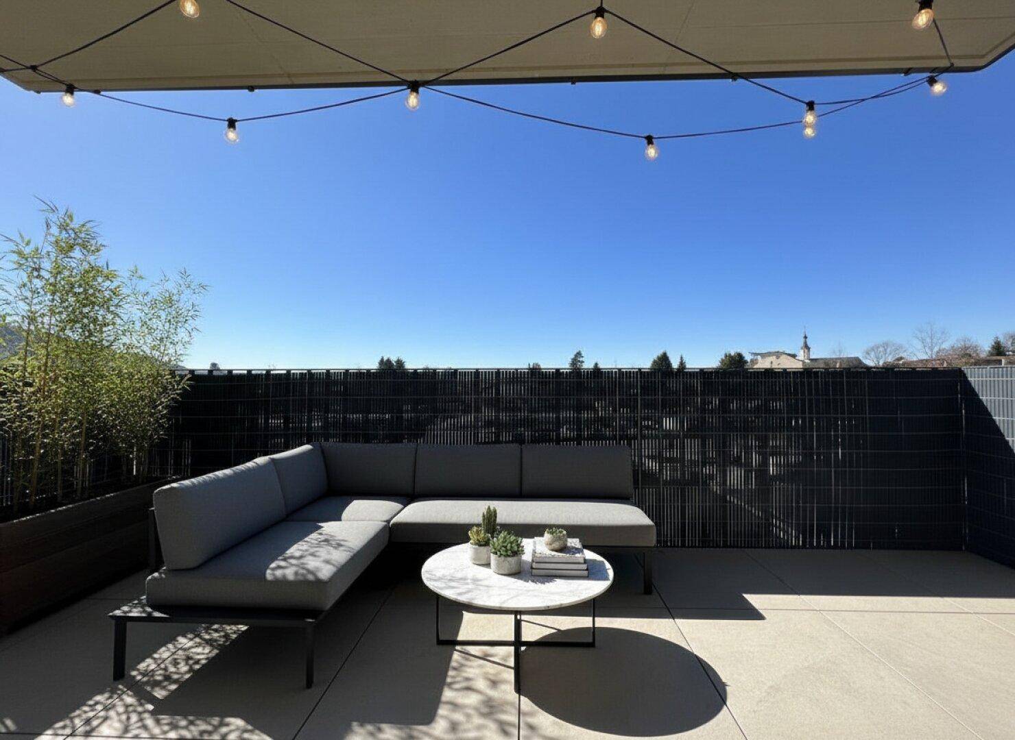Große Dachterrasse mit Lounge-Sofa, Pflanzen und Lichterkette unter blauem Himmel.
