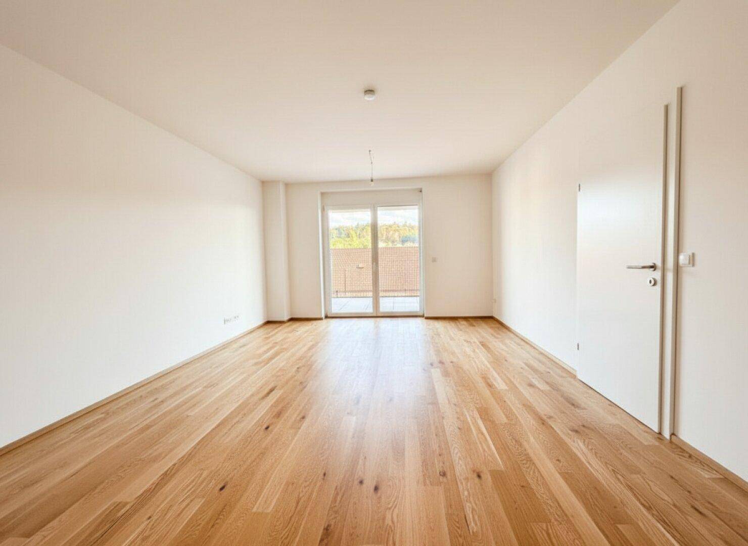 Leeres, helles Zimmer mit Holzboden und Balkonzugang, bereit zur individuellen Gestaltung.