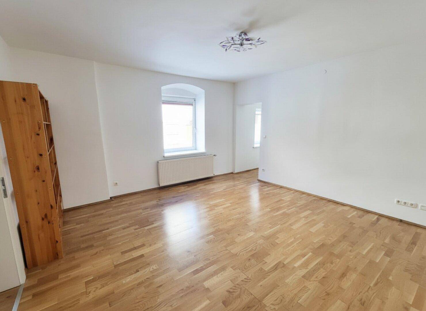 Helles, leeres Zimmer mit Parkettboden und einem Fenster, das viel Tageslicht hereinlässt.