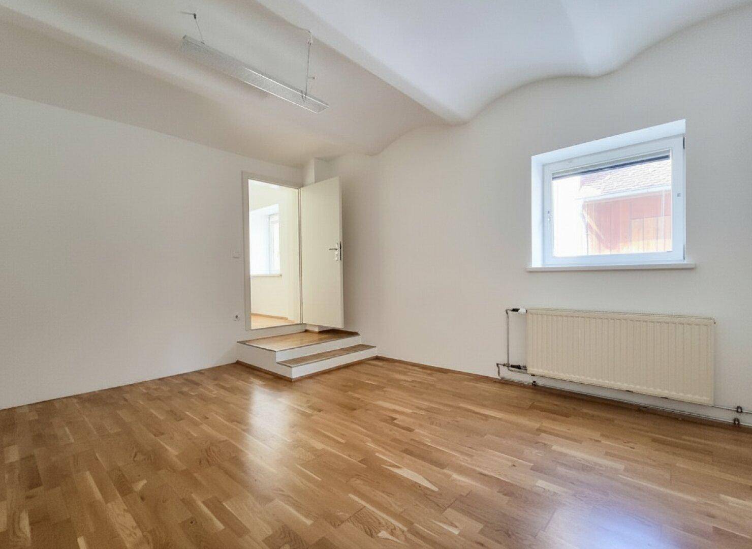Leeres Zimmer mit Parkettboden, hohen Decken und einem Fenster, das für gute Belichtung sorgt.