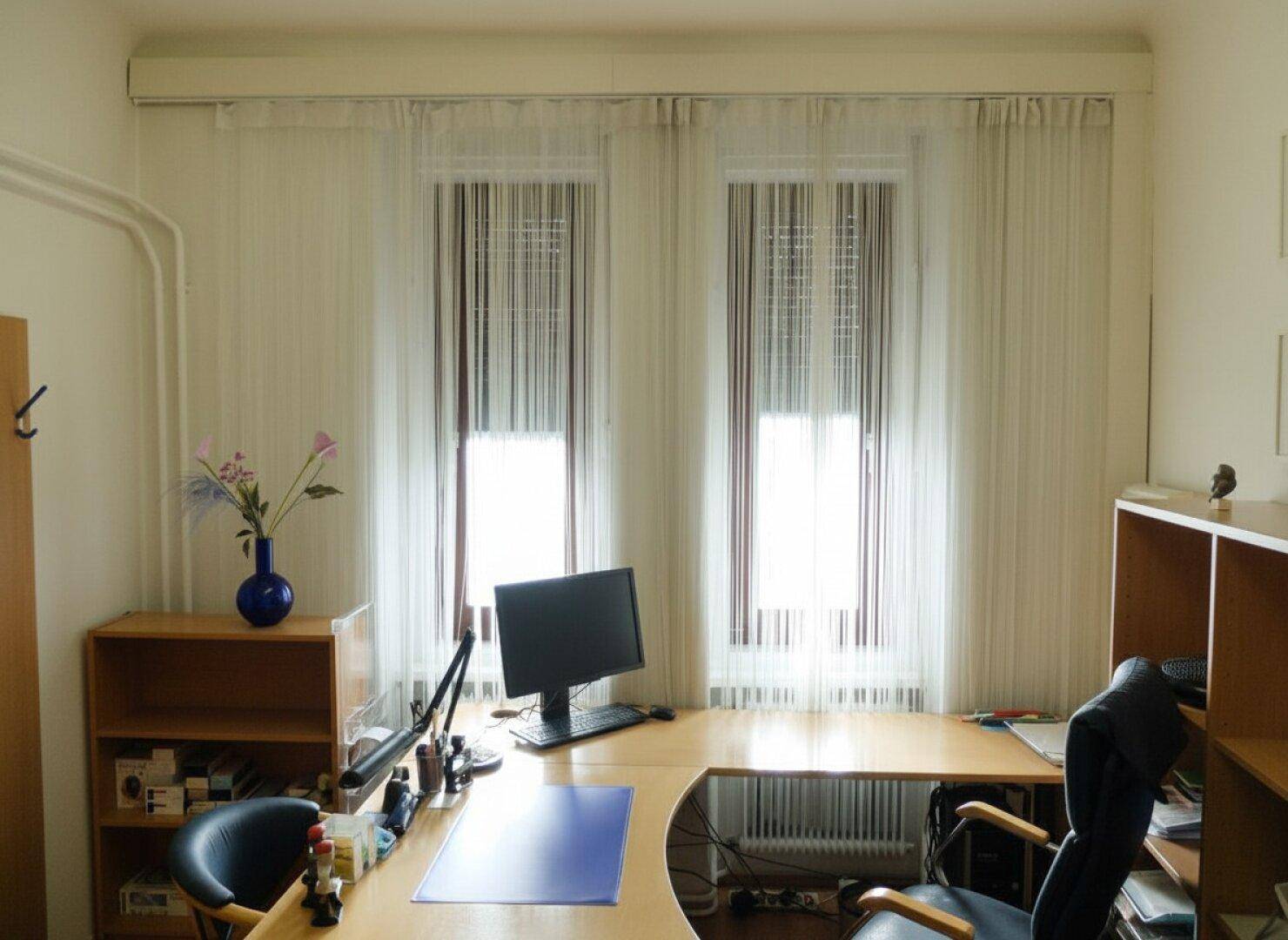 Helles Büro mit zwei Fenstern, Schreibtisch und Regalen für Arbeitsmaterialien.