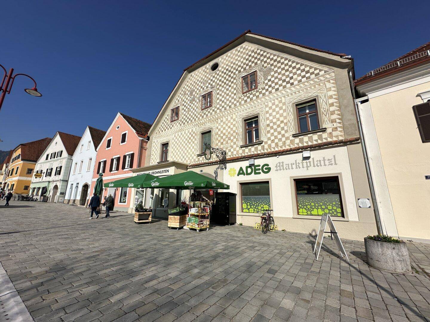 Historische Gebäudezeile mit dekorierter Fassade und belebtem Marktplatz unter blauem Himmel.