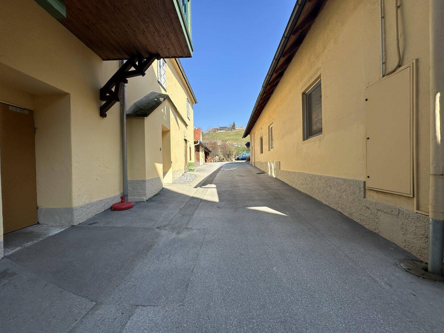 Schmaler Durchgang zwischen Gebäuden mit gelben Fassaden, der zu einem sonnigen Hügel führt.