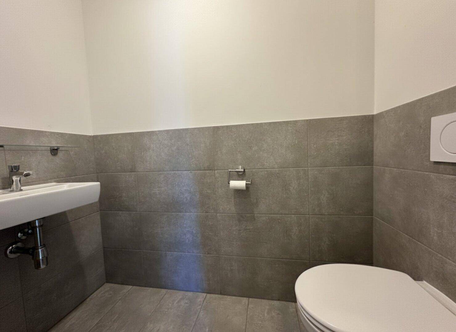 Separates Gäste-WC mit Wand-WC, kleinem Waschbecken und grauen Fliesen.