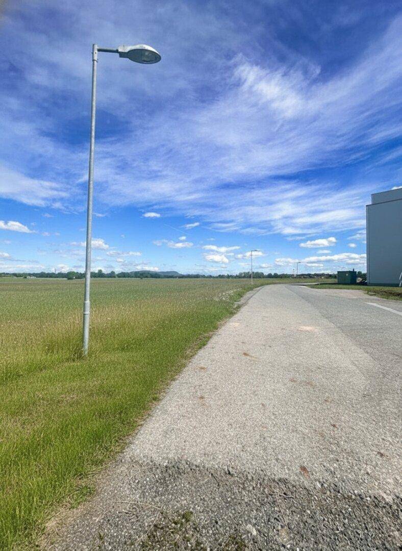Ein Schotterweg führt entlang eines weiten Feldes unter einem bewölkten blauen Himmel mit Laternen und Bergen in der Ferne.