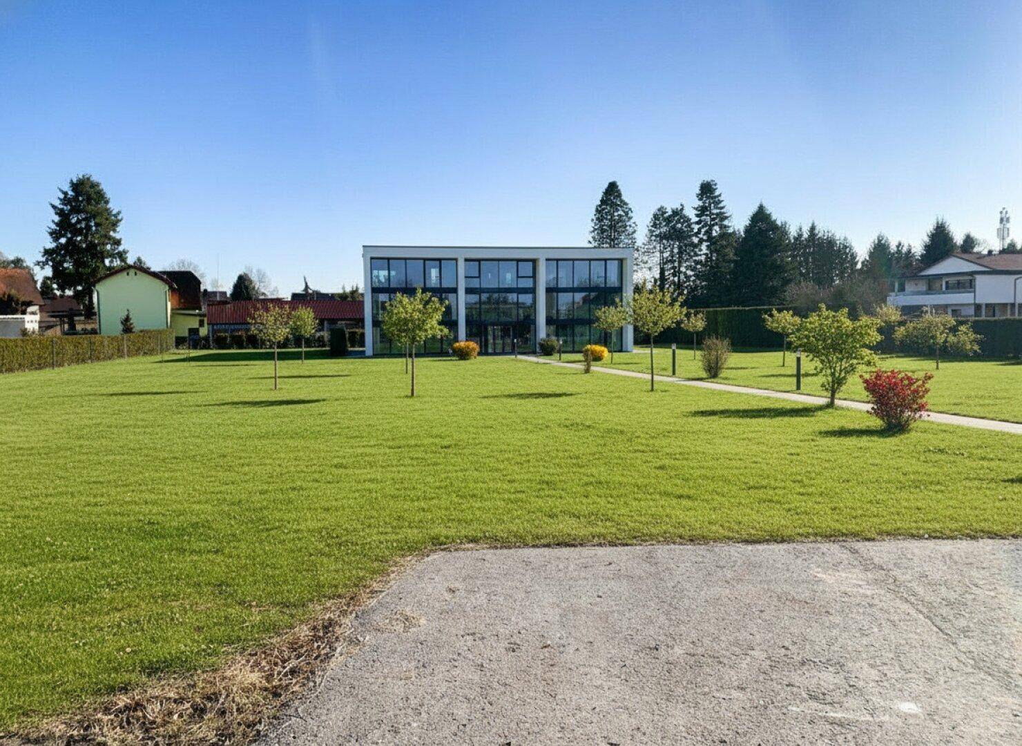 Ein modernes Gebäude mit großen Glasflächen, umgeben von einer gepflegten Grünfläche und jungen Bäumen unter blauem Himmel.