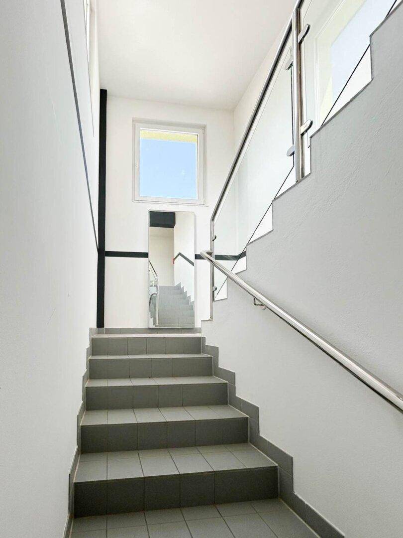 Modernes Treppenhaus mit grauen Fliesenstufen, weißen Wänden und einem Geländer aus Edelstahl und Glas.