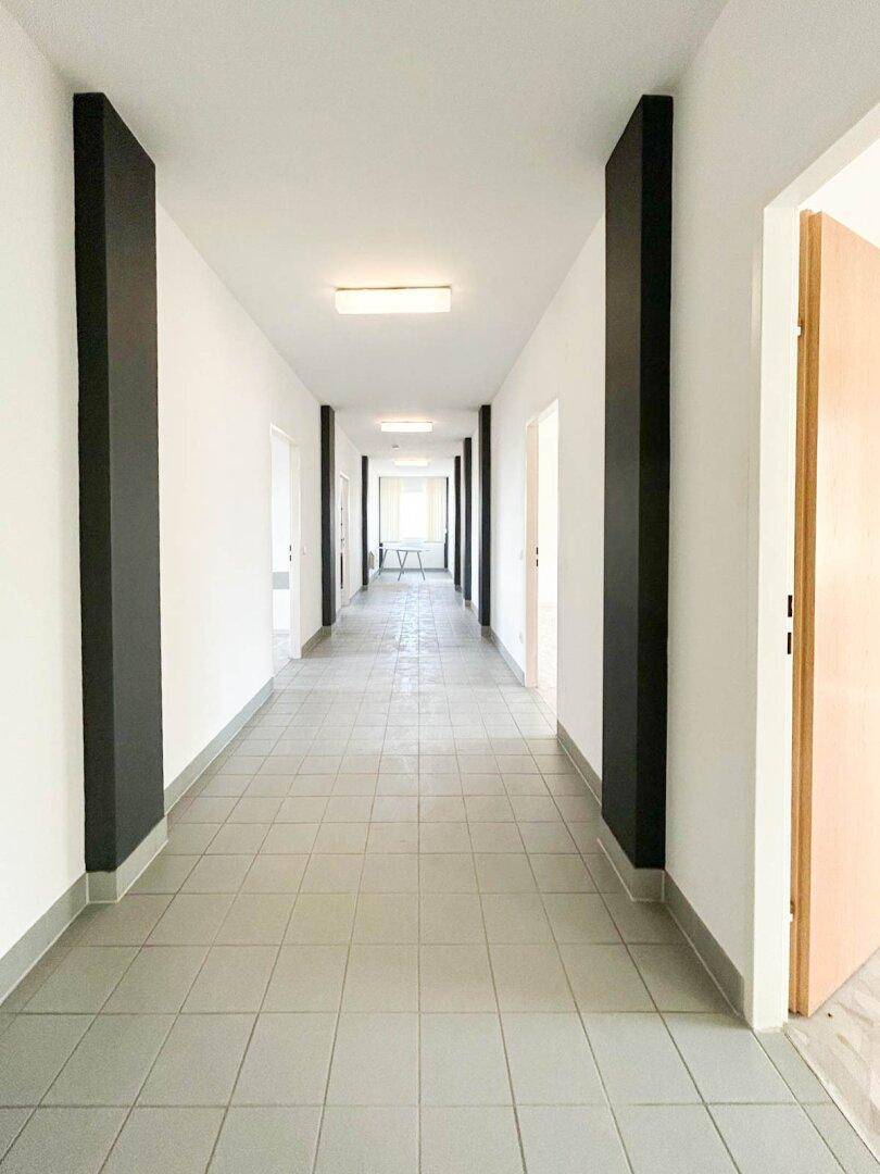 Langer, leerer Flur mit weißen Wänden, grauen Fliesen und schwarzen Akzenten, beleuchtet von Deckenlampen.