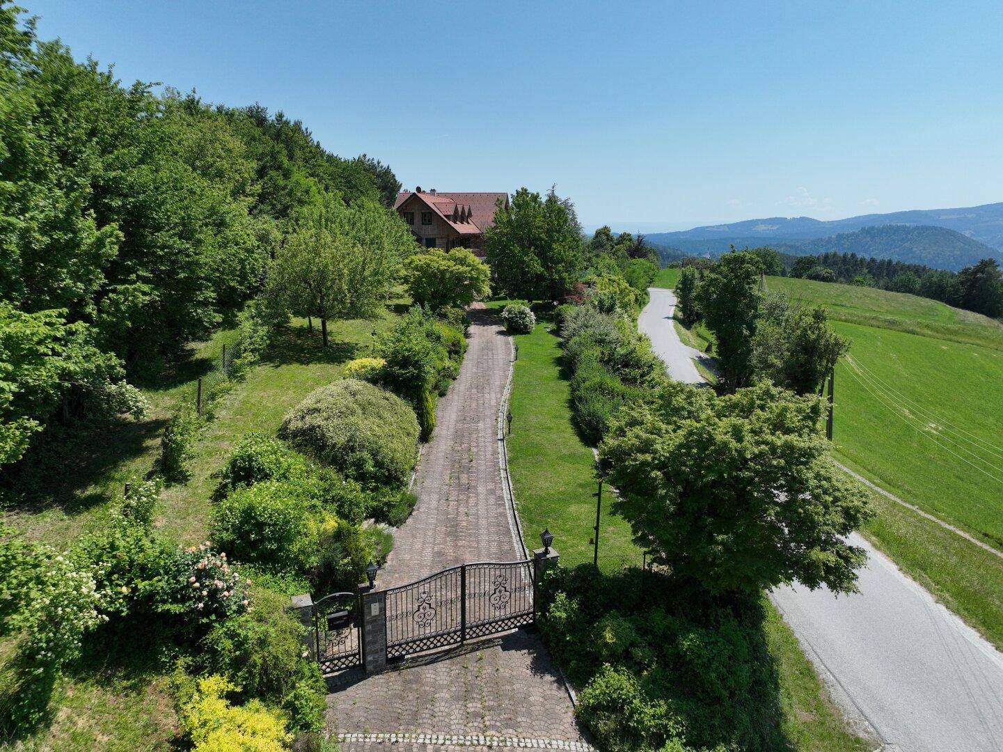 Luftaufnahme des Anwesens mit Zufahrt, großem Tor und dem Haus inmitten grüner Landschaft.