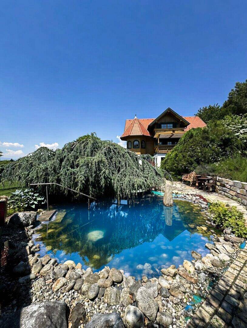 Natürlicher Schwimmteich mit Felsen und einer Hängezeder, im Hintergrund ein traditionelles Holzhaus.