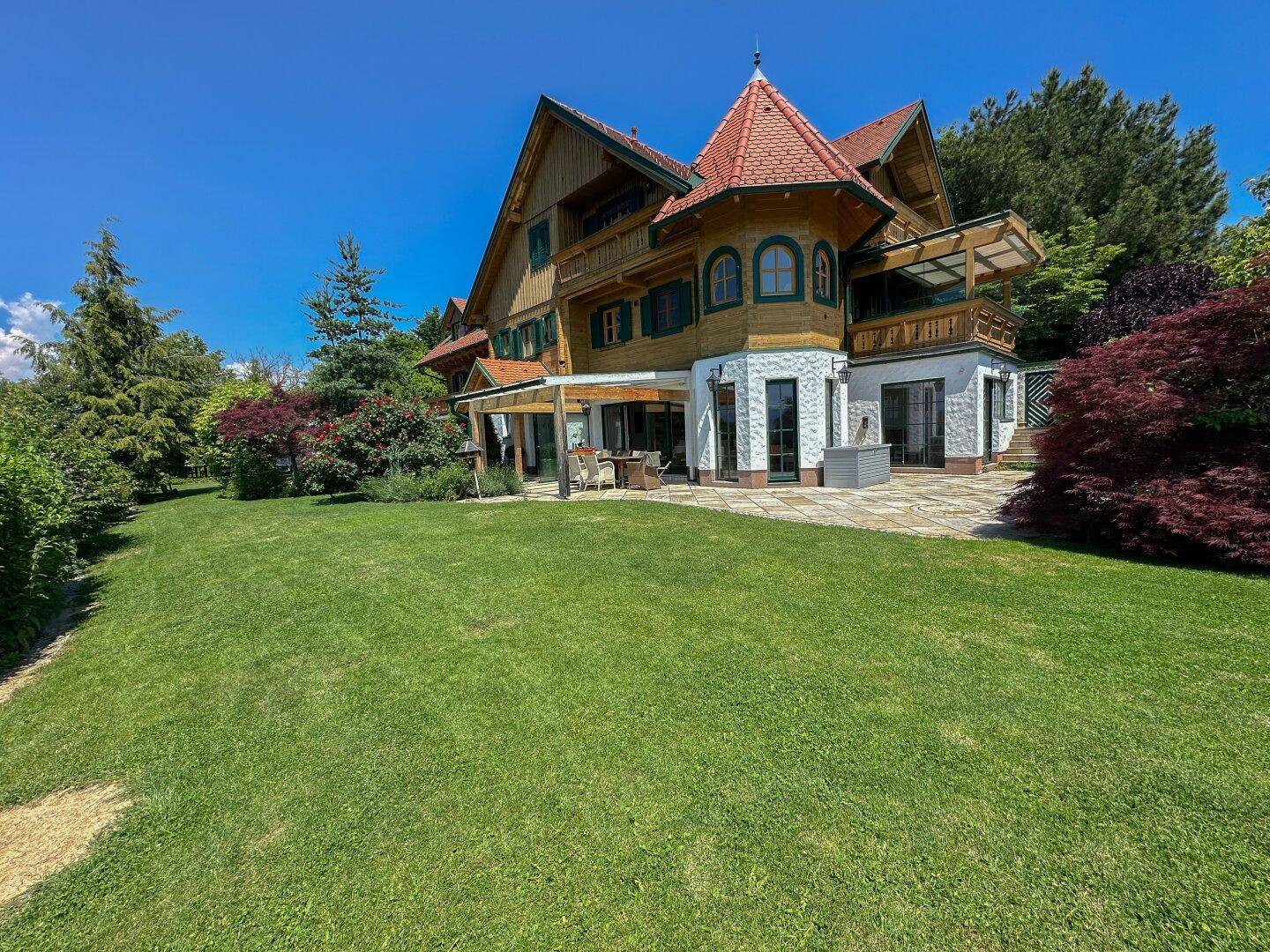 Weitläufiger Rasenbereich vor dem traditionellen Holzhaus mit Terrasse unter blauem Himmel.
