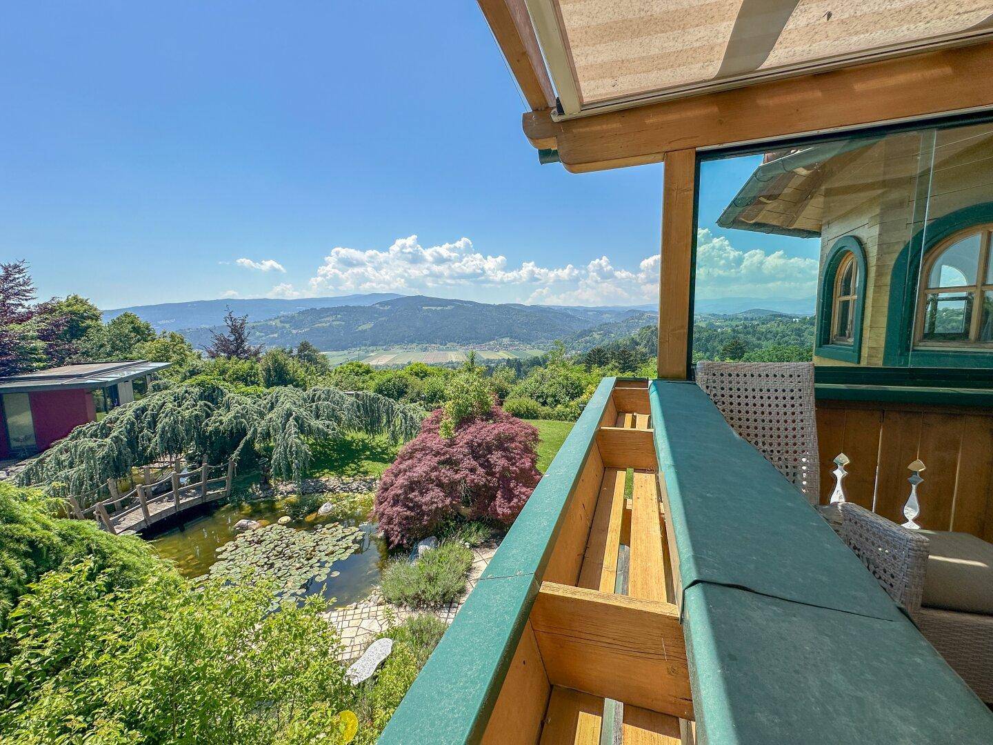 Weitblick vom Balkon über die grüne Landschaft mit Teich und dem charmanten Anwesen.
