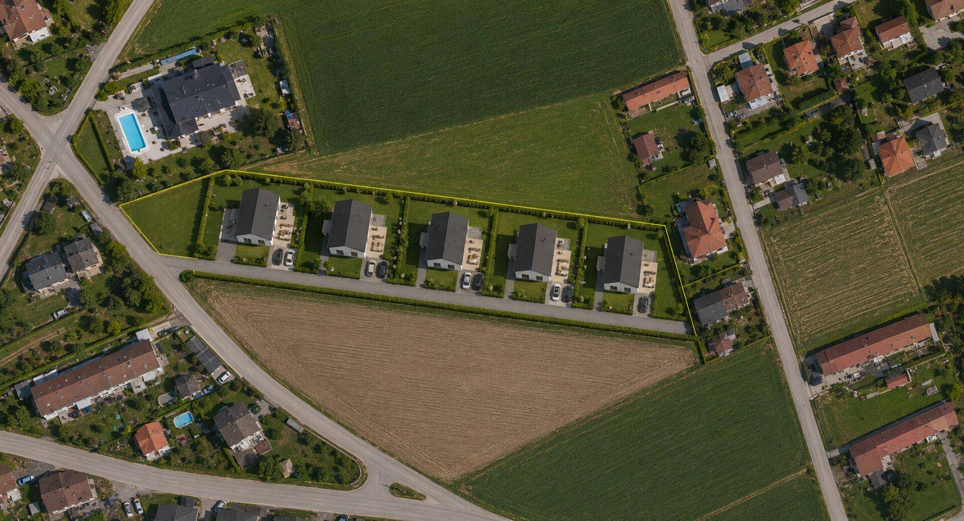Luftaufnahme einer Reihe moderner Neubauten mit Gärten und Parkplätzen, umgeben von grünen Feldern.