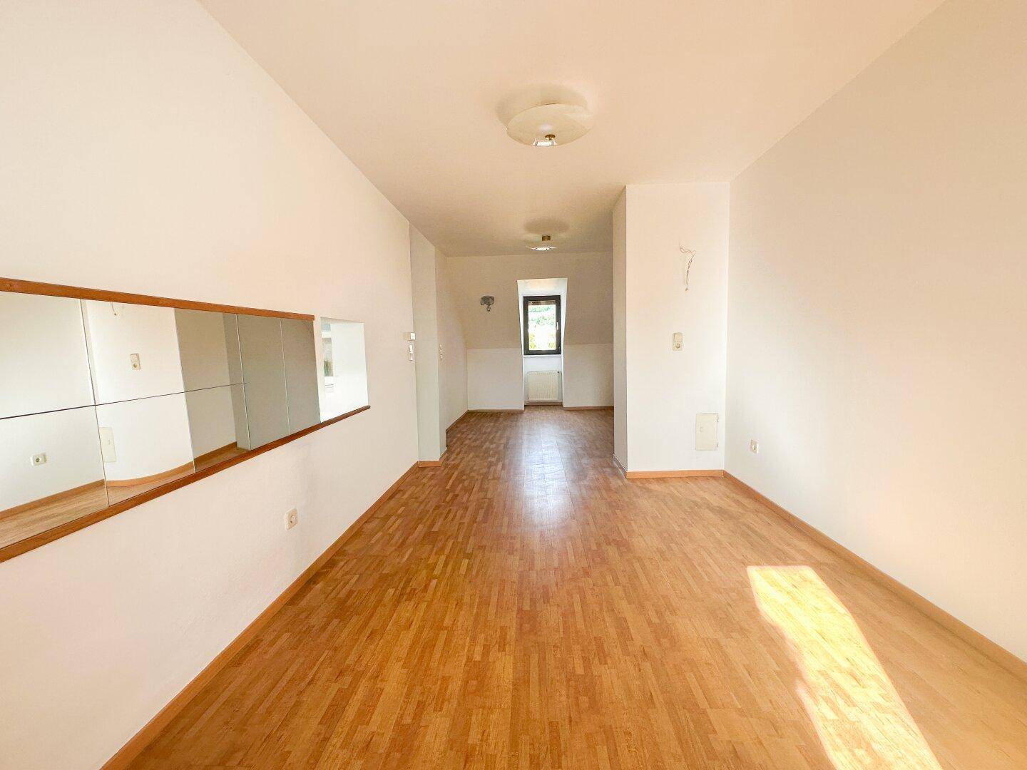 Helles, leeres Zimmer mit Parkettboden und einem Fenster, das viel Tageslicht hereinlässt.