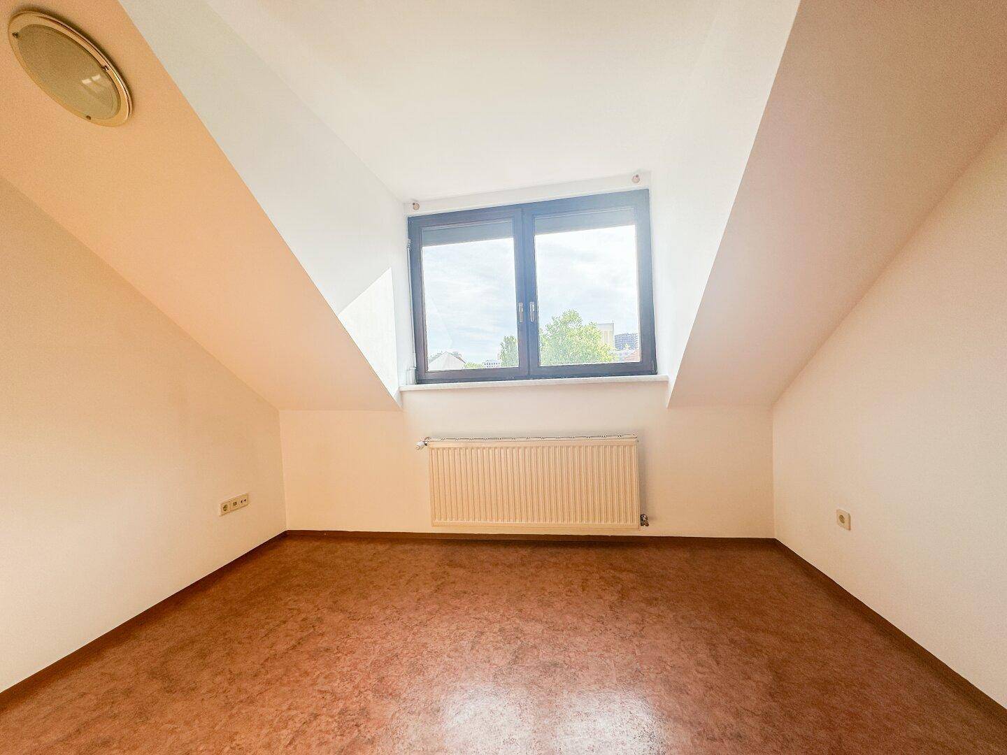 Leeres Schlafzimmer im Dachgeschoss mit Dachschrägen, Fenster und Heizkörper.