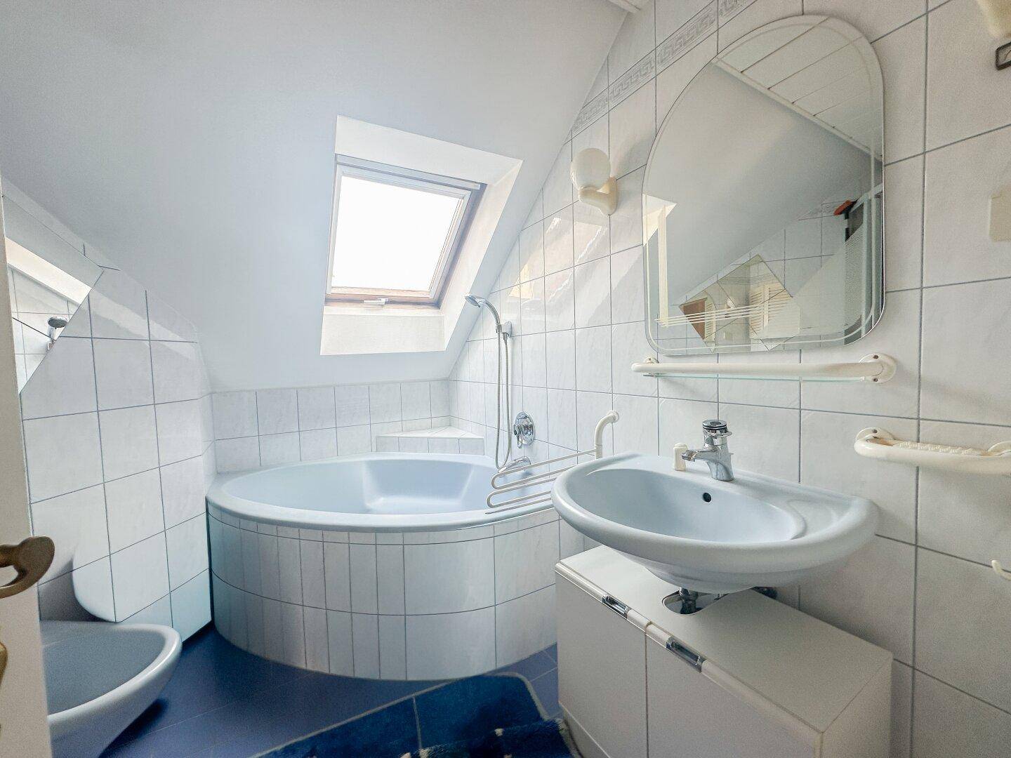 Badezimmer im 80er-Jahre-Stil mit Eckbadewanne, Waschbecken und Dachfenster.