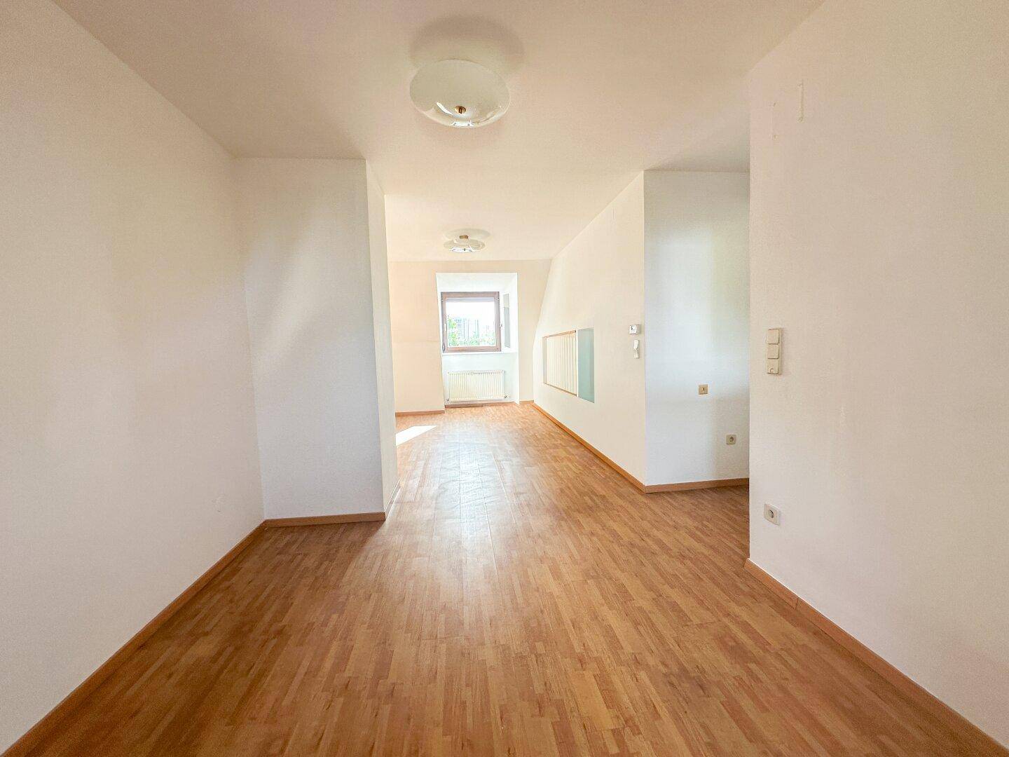 Geräumiges, leeres Zimmer mit Holzboden und einem Fenster, das den Raum hell erscheinen lässt.